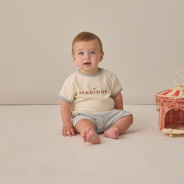 Baby sitting on a plain background wearing a 'Magique' shirt and gray shorts.