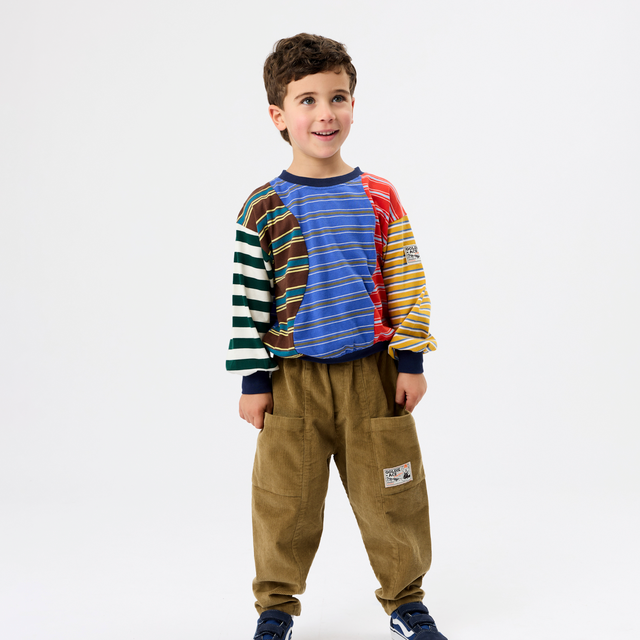 A young boy modeling a colorful long-sleeve knit top with striped and solid color panels, paired with brown pants and navy sneakers, against a plain white studio background.