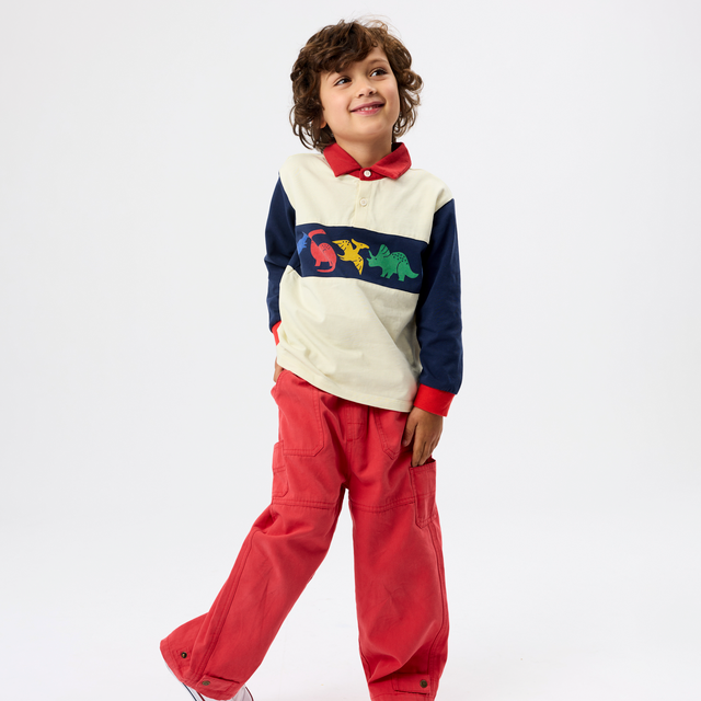 A young boy modeling a color-blocked long-sleeve polo shirt with red pants, standing against a plain white studio background.