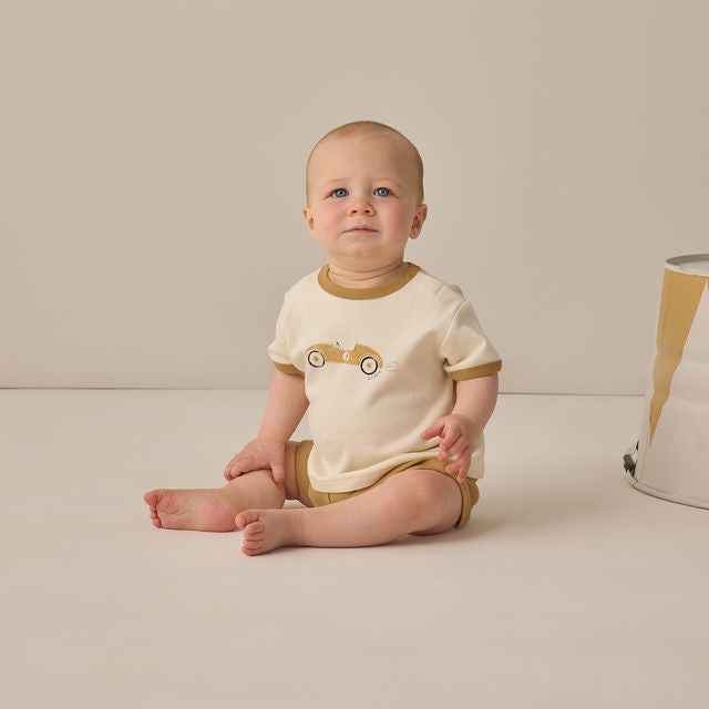 Baby wearing a white and brown outfit with a car design, sitting on a beige surface.