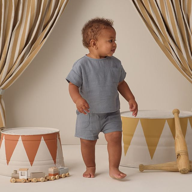 Baby in a blue outfit standing in a room with decorative curtains and toys.