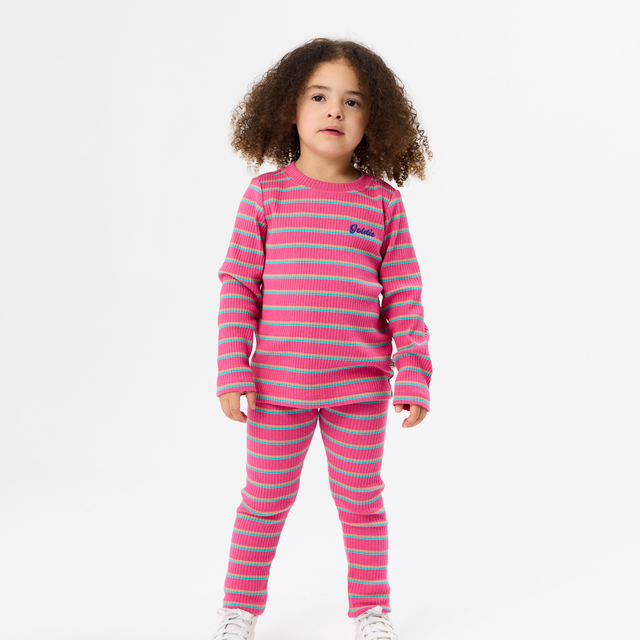 A young girl with curly brown hair stands against a plain white studio background wearing a matching two-piece pajama set with horizontal pink and light blue stripes, featuring a long-sleeve top and pants. She is wearing white sneakers.