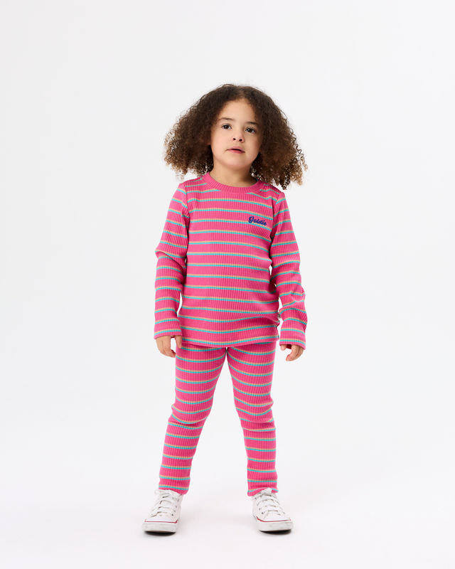 A young girl with curly brown hair stands against a plain white studio background wearing a matching two-piece pajama set with horizontal pink and light blue stripes, featuring a long-sleeve top and pants. She is wearing white sneakers.