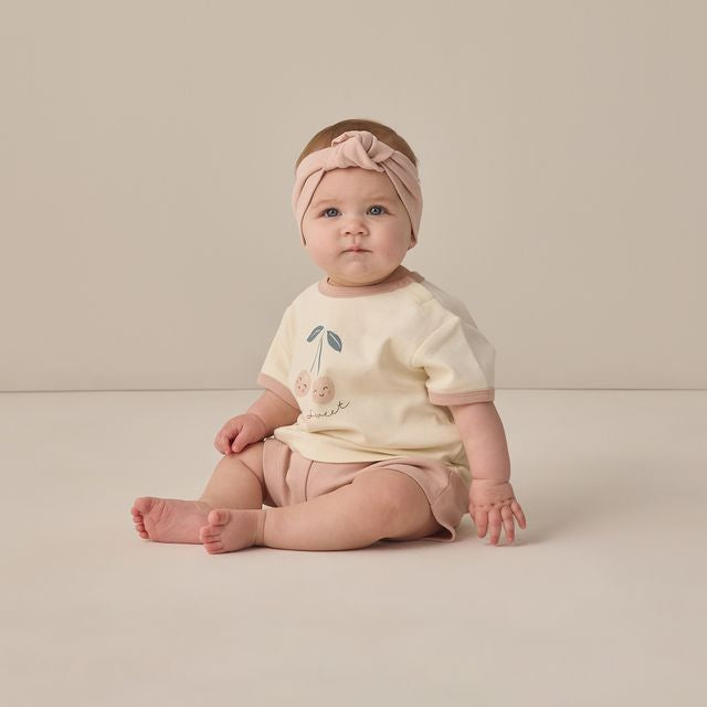 Baby sitting on a plain background wearing a light-colored outfit with a headband.