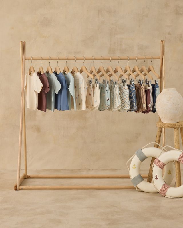 Wooden clothing rack with baby clothes against a beige wall, with decorative items on the floor.