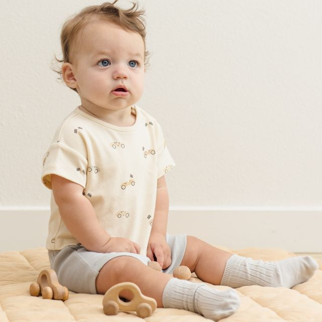 Baby sitting on a beige blanket with wooden toys, wearing a light-colored shirt and gray shorts.
