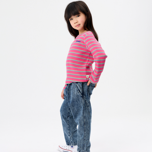 A young girl modeling a pink and blue striped long-sleeve knit top, paired with blue jeans and white sneakers, standing against a plain white studio background.