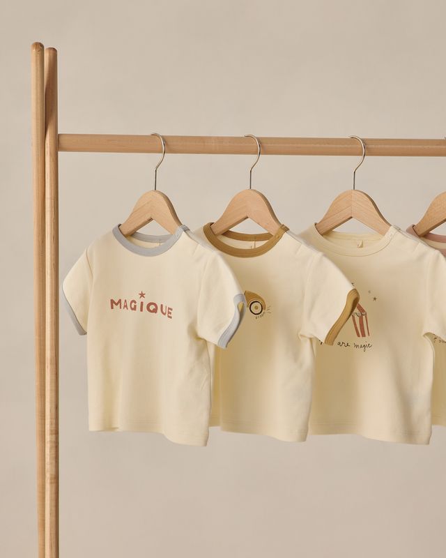 Three children's t-shirts on hangers against a beige wall.