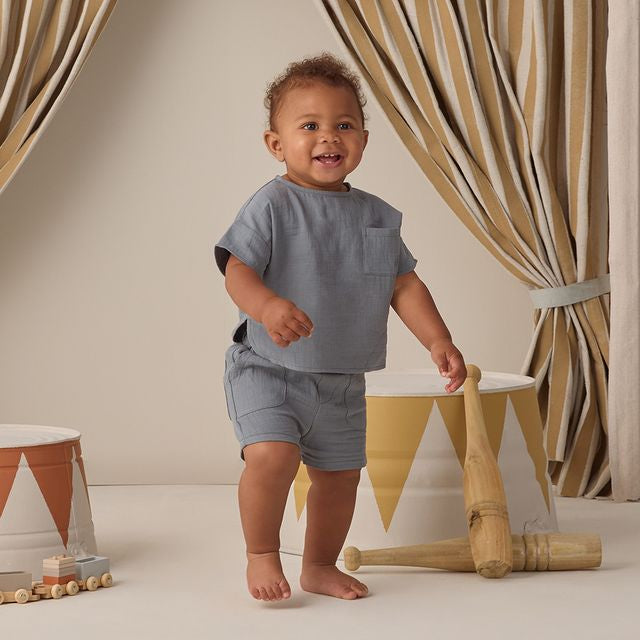 Child in gray outfit standing in a room with striped curtains and wooden toys.