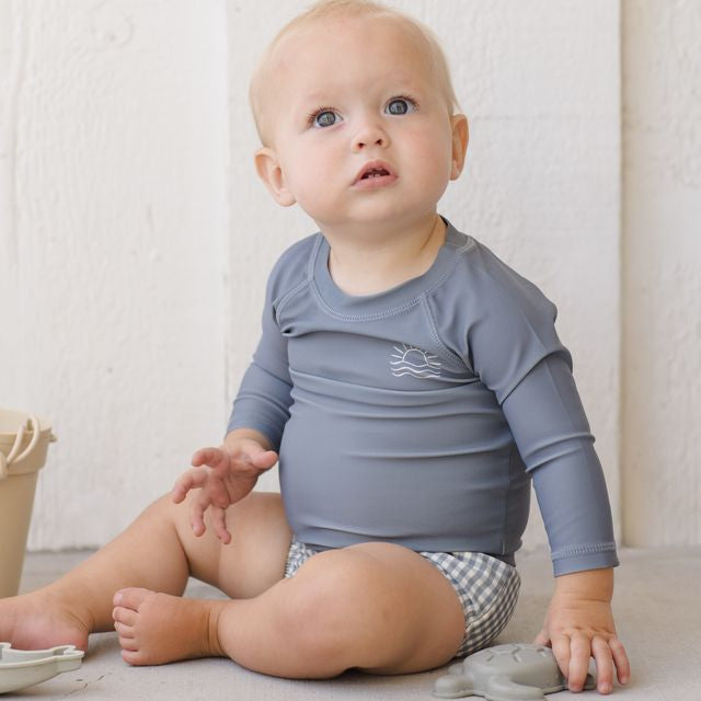Baby wearing a blue long-sleeve shirt and checkered shorts sitting on a light-colored floor.