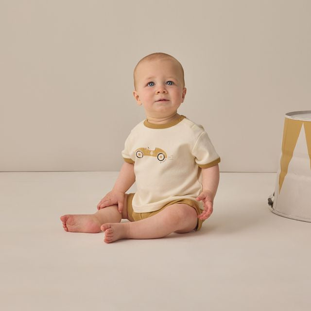 Baby sitting on a beige floor wearing a white and brown outfit with a car design.
