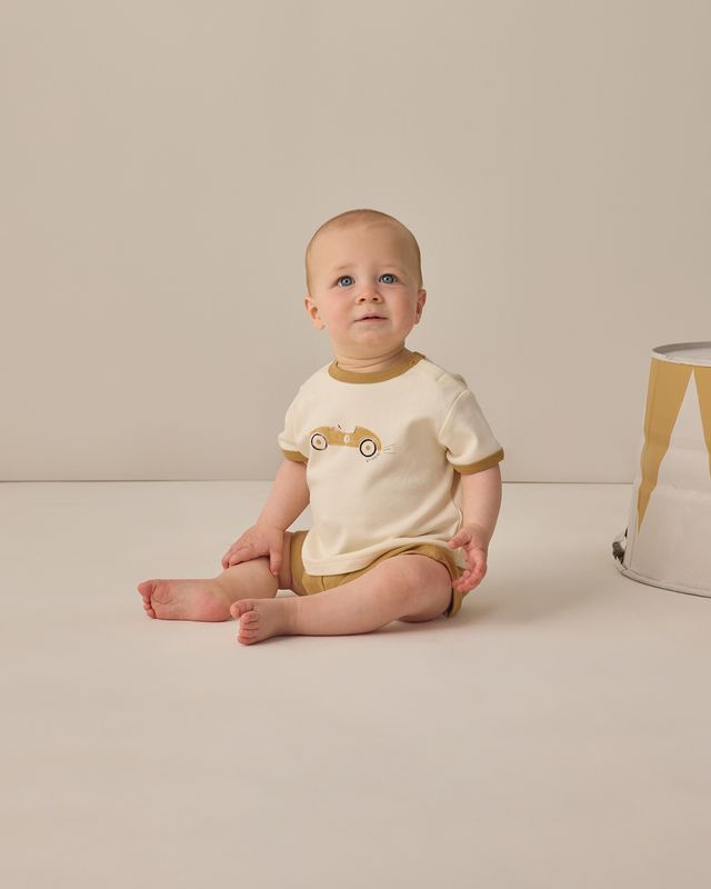 Baby sitting on a beige floor wearing a white and brown outfit with a car design.