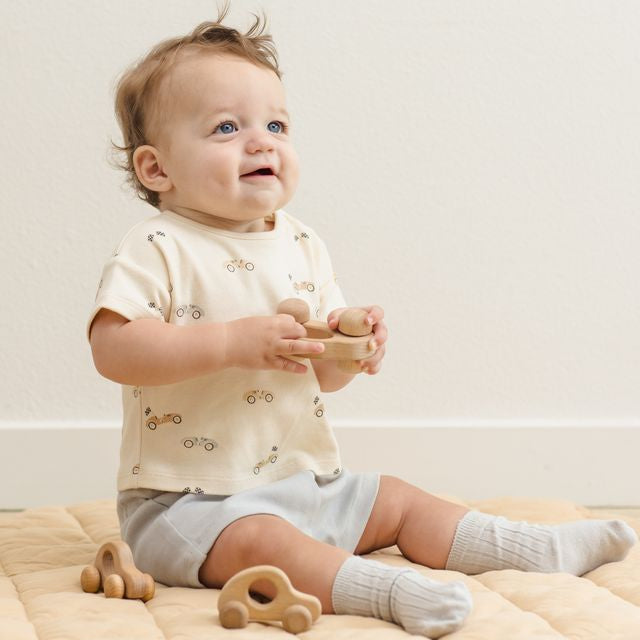 Baby sitting on a beige blanket with wooden toys, wearing a light-colored outfit.