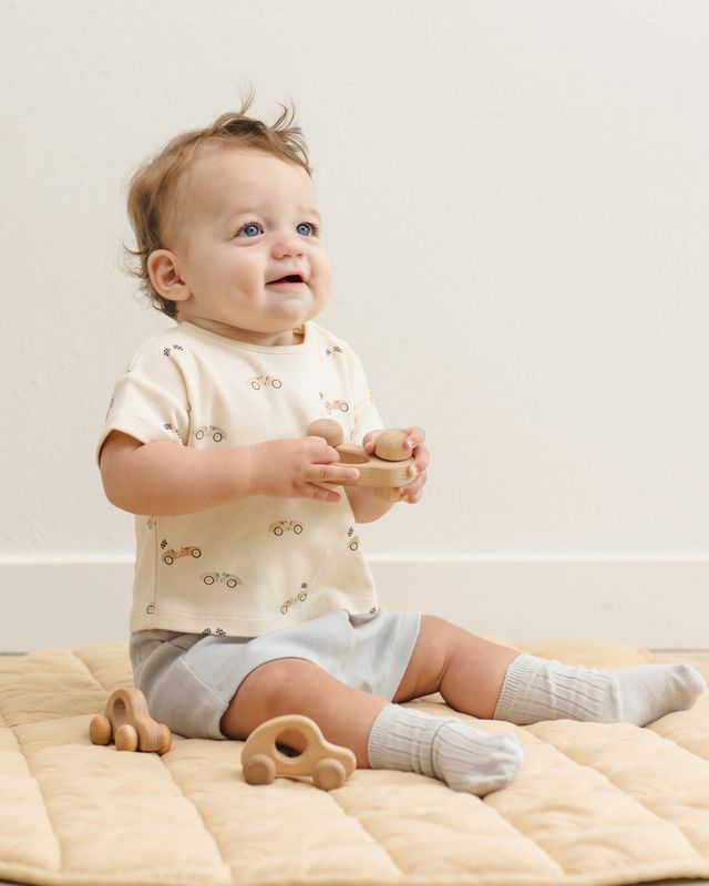 Baby sitting on a beige blanket with wooden toys, wearing a light-colored outfit.