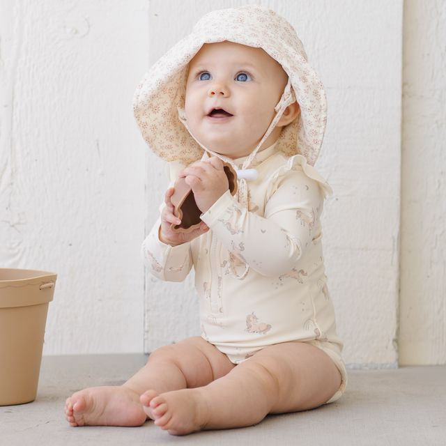 Baby wearing a white hooded outfit sitting on a light-colored surface with a neutral background.