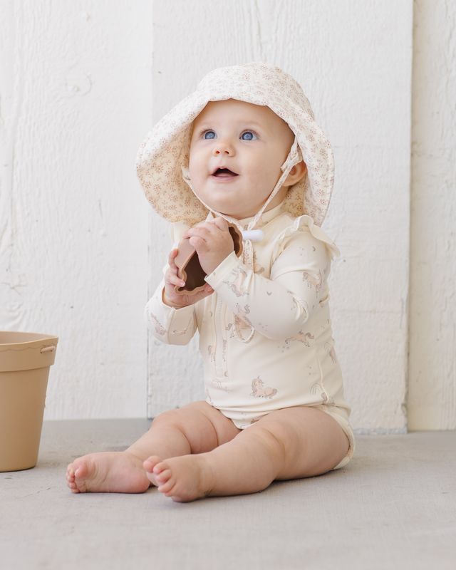 Baby wearing a white hooded outfit sitting on a light-colored surface with a neutral background.