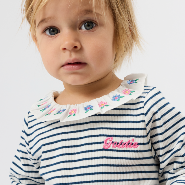 A close-up of a toddler wearing a navy and white striped long-sleeve top with a white ruffled collar decorated with small colorful flowers and a pink embroidered 'Grishie' logo on the chest, against a plain light gray background.