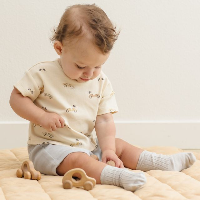 Baby sitting on a beige blanket with wooden toys, wearing a light-colored outfit.