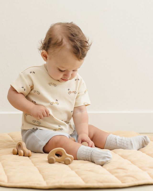 Baby sitting on a beige blanket with wooden toys, wearing a light-colored outfit.