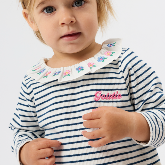 A close-up of a young child wearing a navy and white horizontally striped long-sleeve top with a white embroidered floral collar and a pink 'Gabbie' script on the chest.