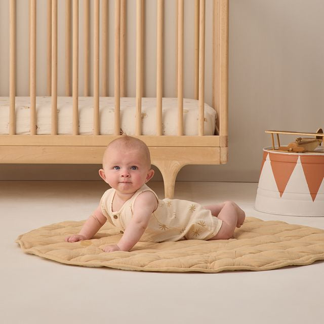 Baby lying on a beige blanket in front of a wooden crib in a nursery.