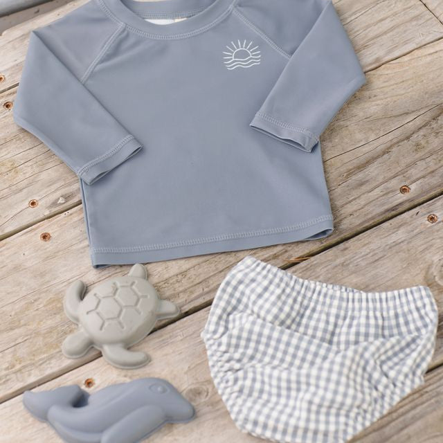 Children's gray long-sleeve shirt with a sun design, checkered shorts, and turtle toys on a wooden surface.