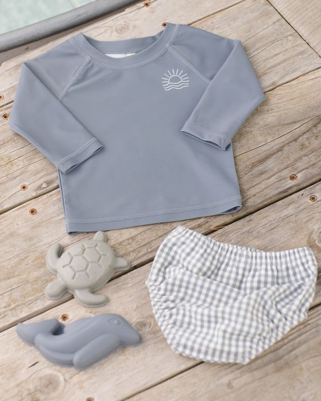 Children's gray long-sleeve shirt with a sun design, checkered shorts, and turtle toys on a wooden surface.