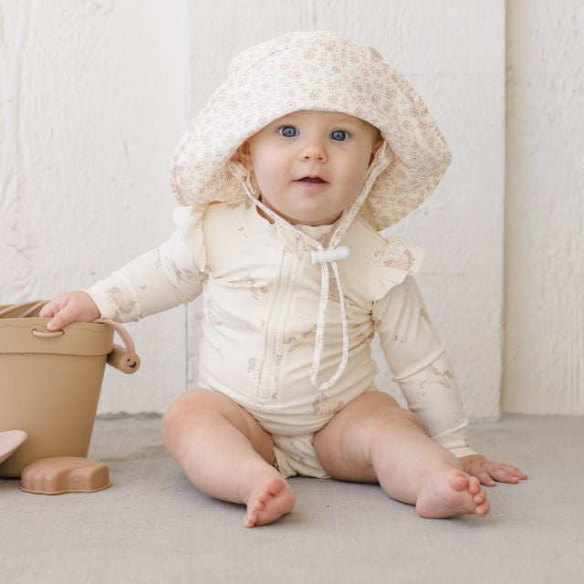 Baby in a cream-colored outfit with a hood sitting on a light surface.
