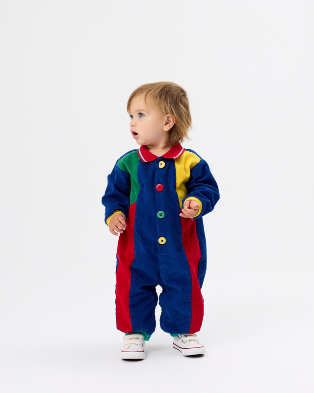 A toddler standing against a plain white studio background, wearing a colorful patchwork-style one-piece outfit with blue, red, green, and yellow sections and large decorative buttons down the front.