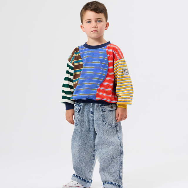 A young boy stands against a light gray studio background wearing a colorful striped long-sleeve shirt with blue jeans and white sneakers.