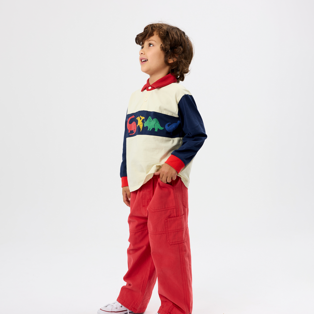 A young boy wearing a color-blocked long-sleeve sweater with dinosaur graphics, red cargo pants, and white sneakers, standing in profile against a plain white studio background.