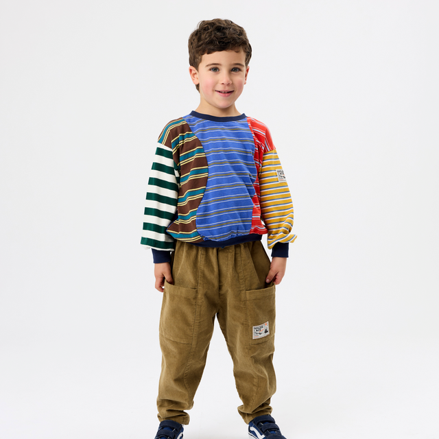 A young boy modeling a colorful long-sleeve shirt with striped panels on the sleeves and chest, paired with brown pants and navy sneakers, against a plain white studio background.