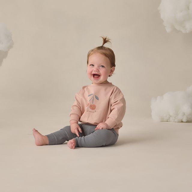 Child wearing a pink sweatshirt with a cherry design, sitting on a beige floor with white cloud decorations.