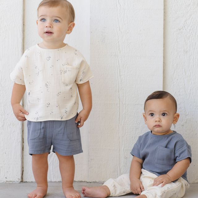 Two toddlers wearing light-colored clothing against a white wooden panel background