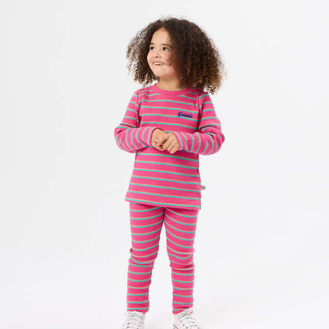 A young girl with curly brown hair stands against a white studio background wearing a matching pink and light blue striped long-sleeve top and pants set, paired with white sneakers.