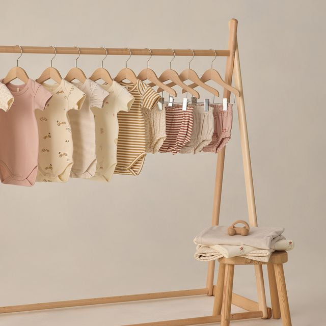Row of baby rompers on a wooden rack with a stool underneath against a beige background
