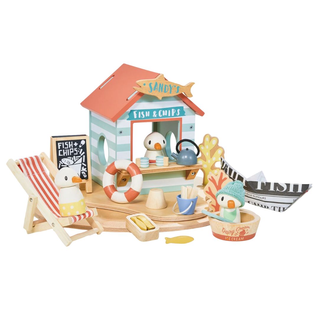 A children's wooden beach bar and boat playset with a striped lounge chair, small boat, toy fish, play money, and a fish & chips counter with toy ducks, set on a round wooden base.