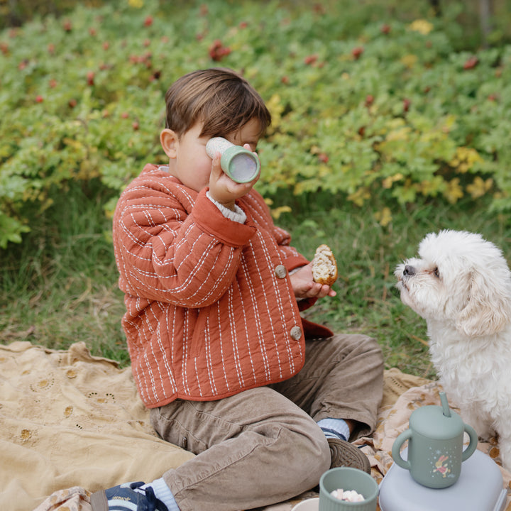 A young child wearing an orange quilted jacket and brown pants sitting on a picnic blanket outdoors, holding a pastry in one hand while looking through a Mushie kaleidoscope, with a small white dog sitting nearby.