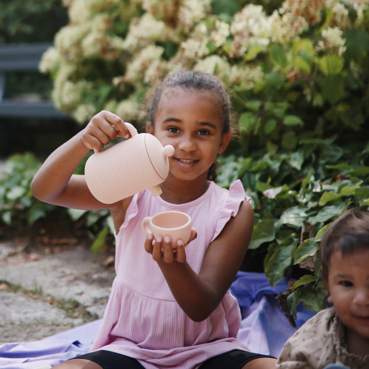 A young child wearing a blue floral print outfit and a brown beanie sits on a curved pink wooden bench outdoors, holding a small toy teapot, while another person tends to a baby nearby.