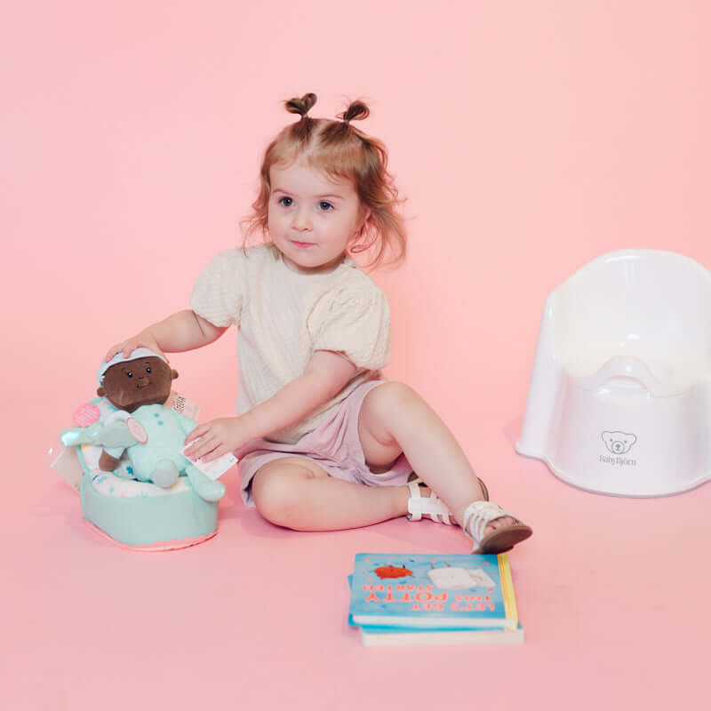 A toddler sits on a pink studio floor wearing a cream short-sleeve top and pale pink shorts, holding a small doll in a light blue outfit. A white baby potty chair is visible to the right, and a colorful book lies in front.