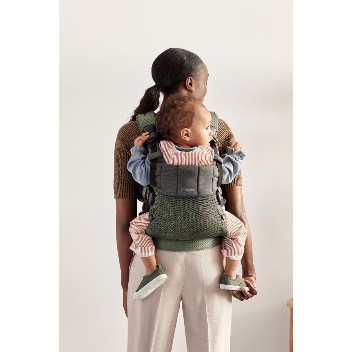 Woman carrying a child in a green baby carrier against a plain background