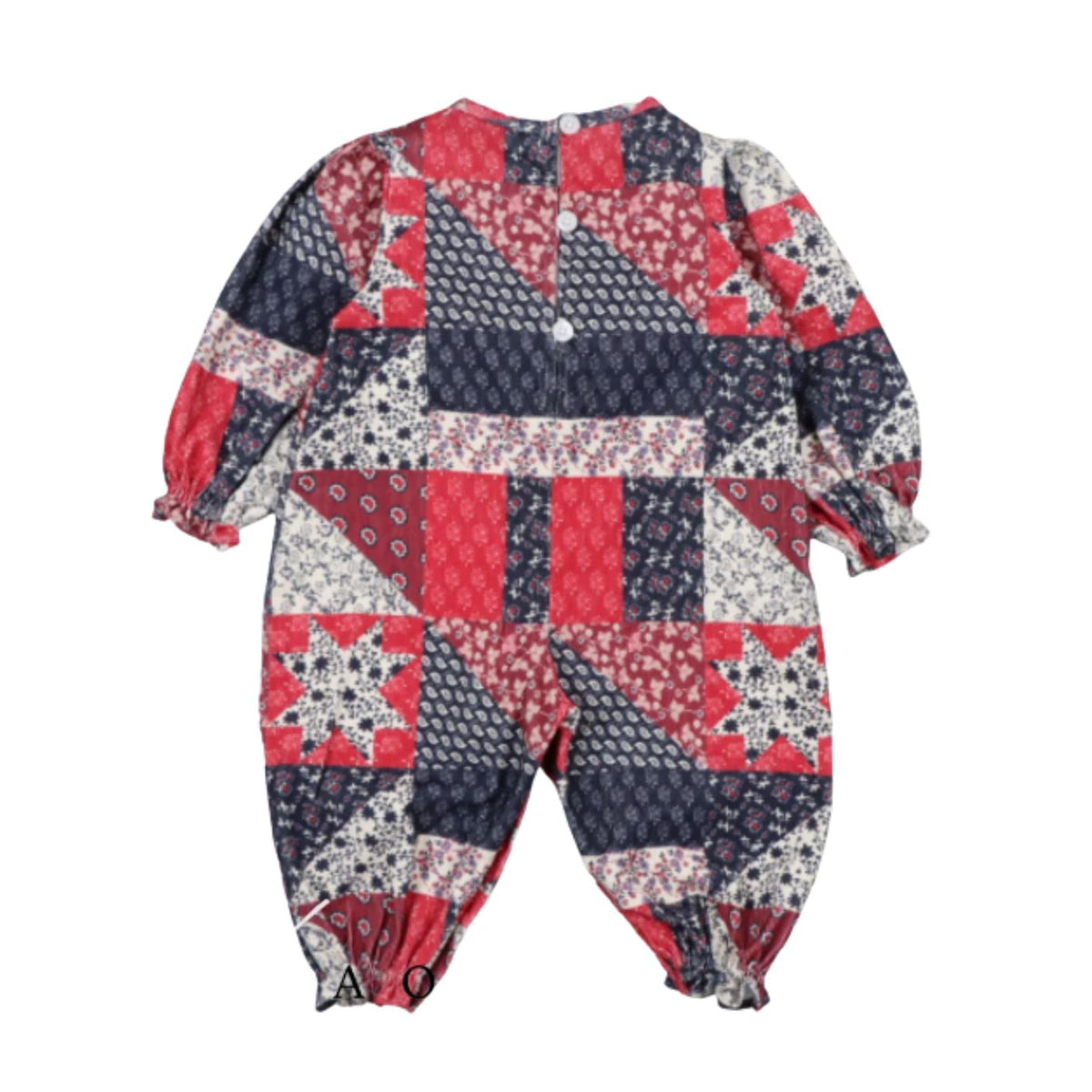 A back-view photo of the Quill Baby Jumpsuit by The New Society against a white background. The one-piece long-sleeve garment features a vintage-inspired patchwork quilt pattern in red, navy blue, and white, with back snap closures and gathered elastic cuffs at the wrists and ankles.