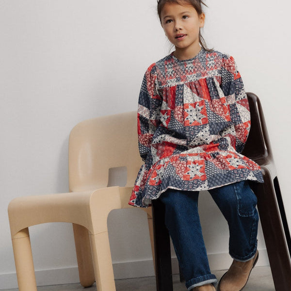 A young child sitting on a black chair next to a light-colored chair in a minimalist room, wearing The Quill Dress by The New Society with dark pants and boots.