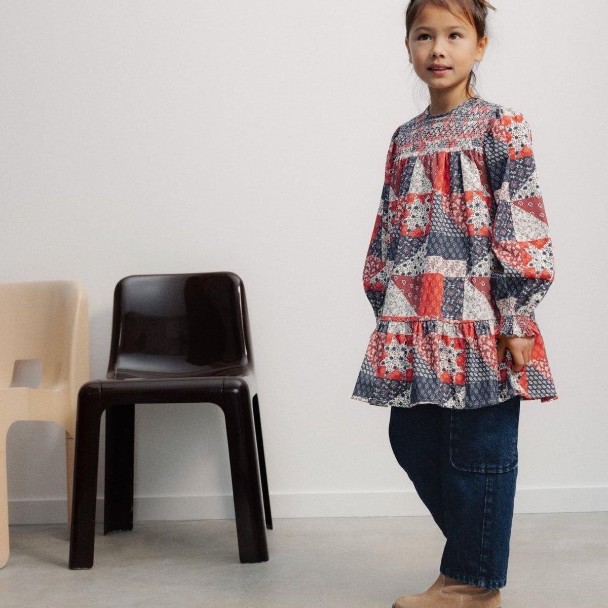 A young child standing and wearing The Quill Dress by The New Society with dark pants and boots, next to a black chair in a minimalist room.
