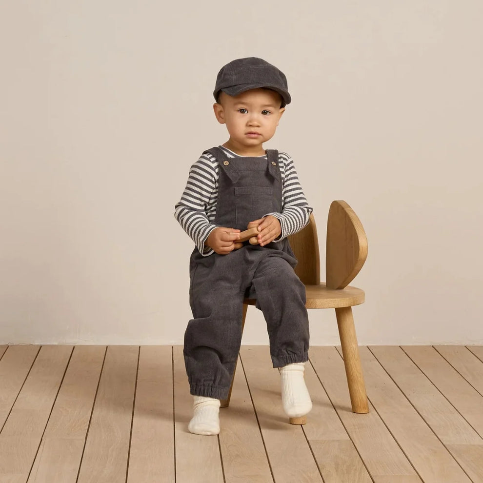 A toddler wearing a dark gray cap, a black and white striped long-sleeve shirt, and dark gray overall pants sits on a small wooden chair against a warm beige background.