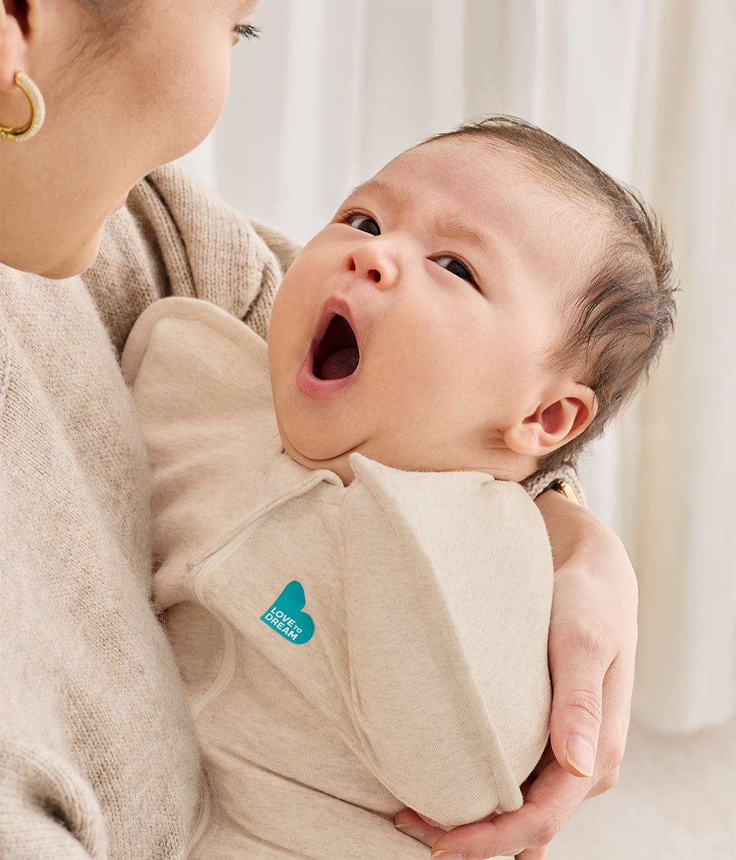 Baby wearing a beige wrap with a visible brand logo, held by an adult against a neutral background
