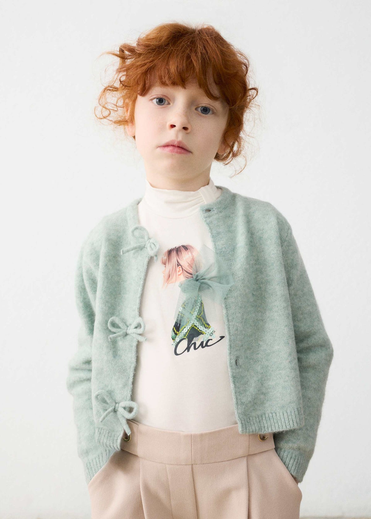 Child wearing a light green cardigan over a white shirt with floral design and beige pants on a white background