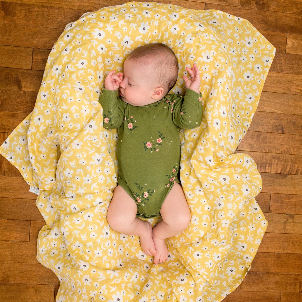 Baby in a green onesie lying on a yellow floral blanket on a wooden floor