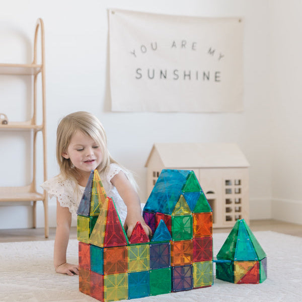 A young child playing on a rug, building castle structures with colorful translucent Magna-Tiles while a toy house sits in the background.