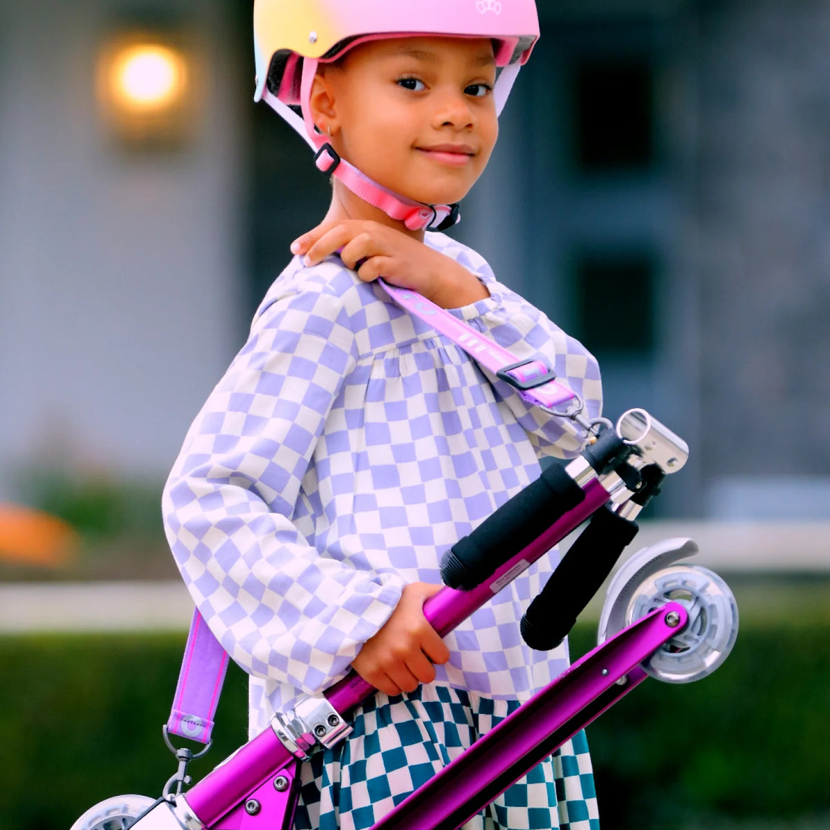 Child wearing a pink helmet and holding a purple scooter outdoors.